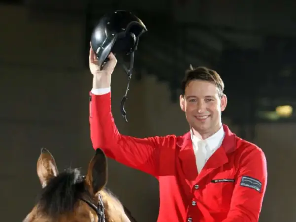 Reiter Franke Sloothaak hält stolz seinen Helm hoch, trägt eine rote Reitjacke und sitzt auf einem braunen Pferd.
